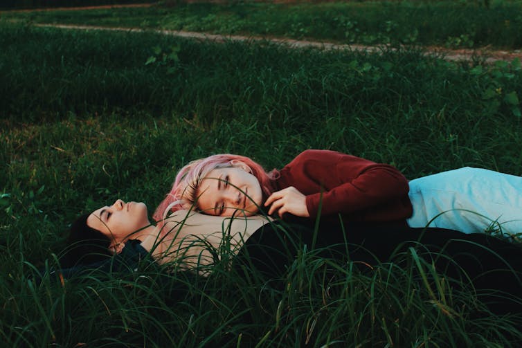 Two teenaged girls lay among grass, cuddling.