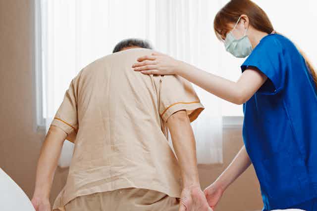 Young woman wearing a surgical mask helps an elderly man get out of bed