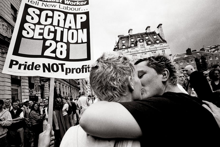 Black and white photo of a lesbian couple kissing at a march, one of the women holds a sign reading Tell New Labour, scrap section 28, pride not profit