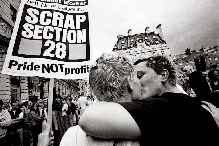 Black and white photo of a lesbian couple kissing at a march, one of the women holds a sign reading Tell New Labour, scrap section 28, pride not profit