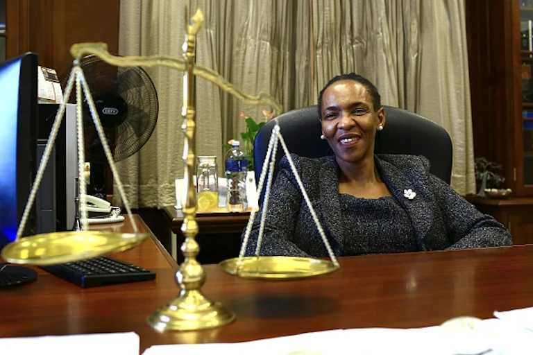 A woman smiles while sitting behind a desk on which lies the scales of justice