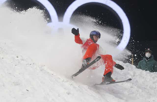 An athlete in orange overalls and on ski crashes into snow with the Olympic logo behind him.