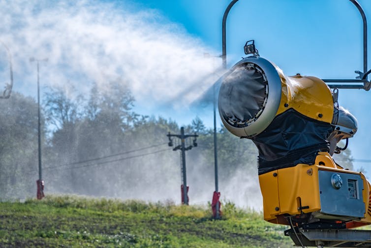 A large yellow machine blasting snow surrounded by mountain terrain during the summer.
