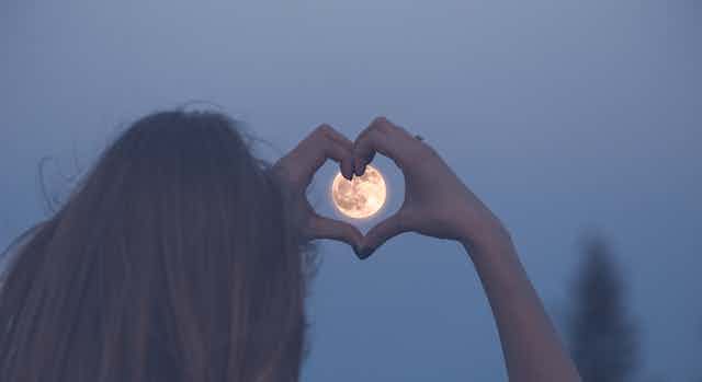 Una mujer enmarca una luna llena con sus manos formando un corazón.