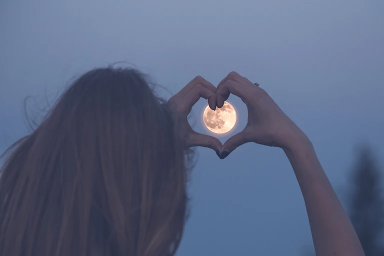 Una mujer enmarca una luna llena con sus manos formando un corazón.