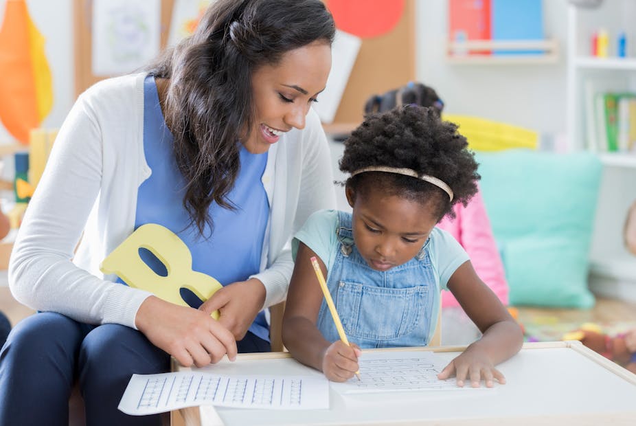 A child care worker and preschool-age girl sit at a table together in a day care classroom and work on handwriting together.