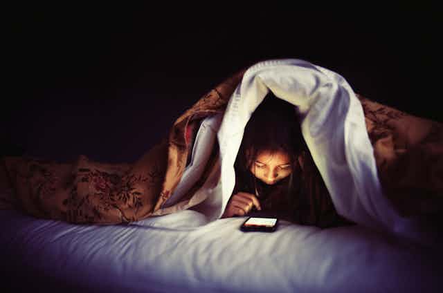 a girl in bed with the covers pulled over her head looks down at a smart phone screen