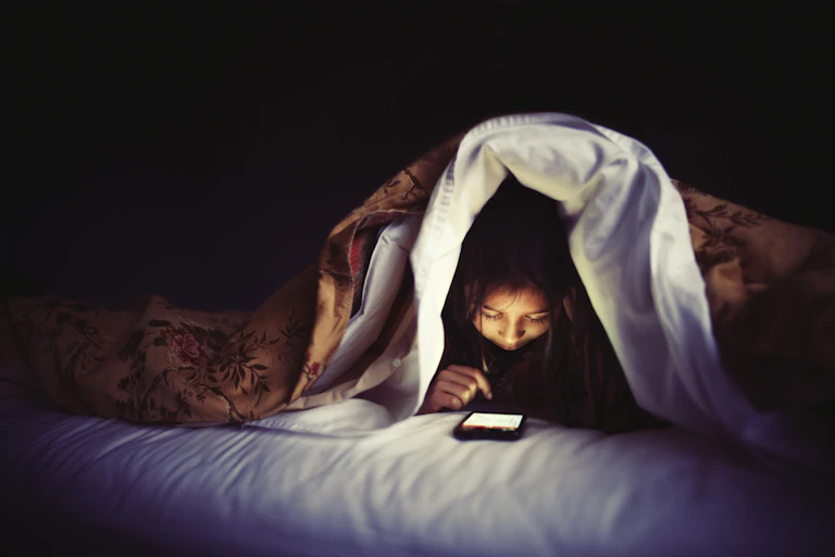 a girl in bed with the covers pulled over her head looks down at a smart phone screen