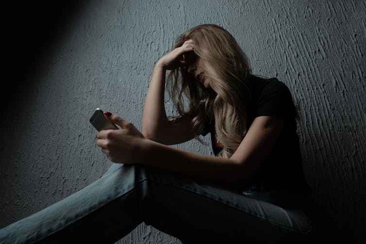A distressed-looking young woman sits in the dark with her head resting on her hand and holding a mobile phone in her other hand