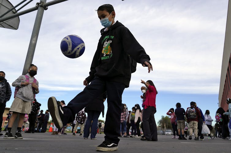 A child wearing a mask kicks a soccer ball in the air as other kids stand nearby