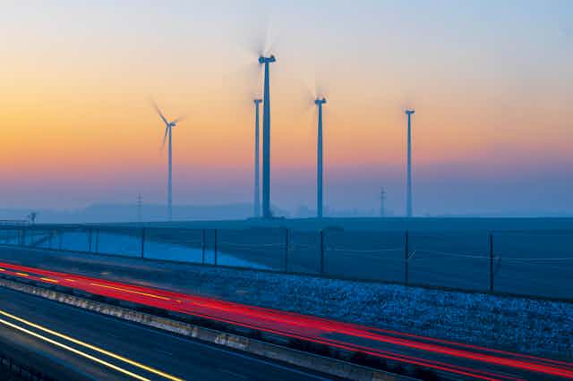 Wind turbines next to a roadway with taillights at sunset.