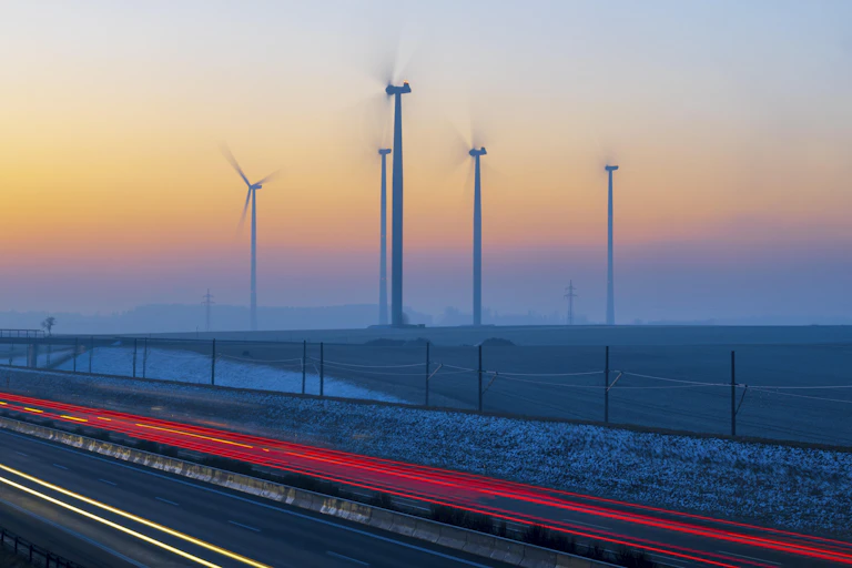 Wind turbines next to a roadway with taillights at sunset.