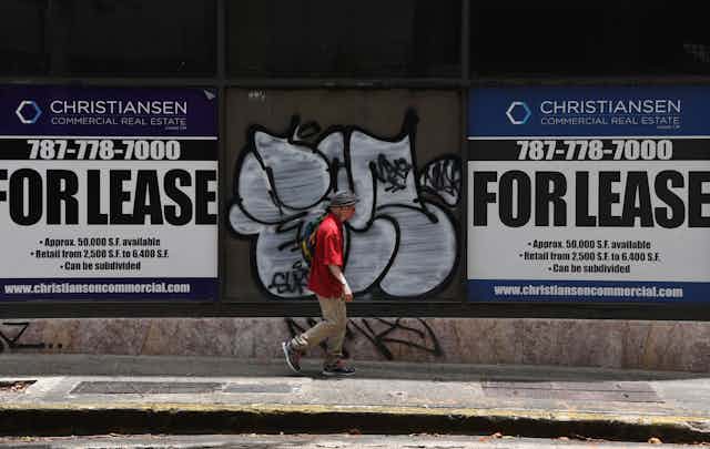 A man wearing a red shirt walks in between two signs that say "FOR LEASE." He walks in front of graffiti on a a city street 