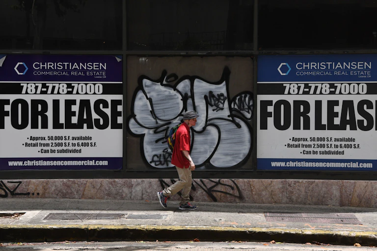 A man wearing a red shirt walks in between two signs that say "FOR LEASE." He walks in front of graffiti on a a city street