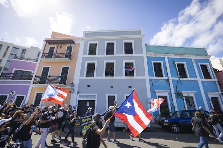 People waving Puerto Rican flags march together past colorful buildings in San Juan