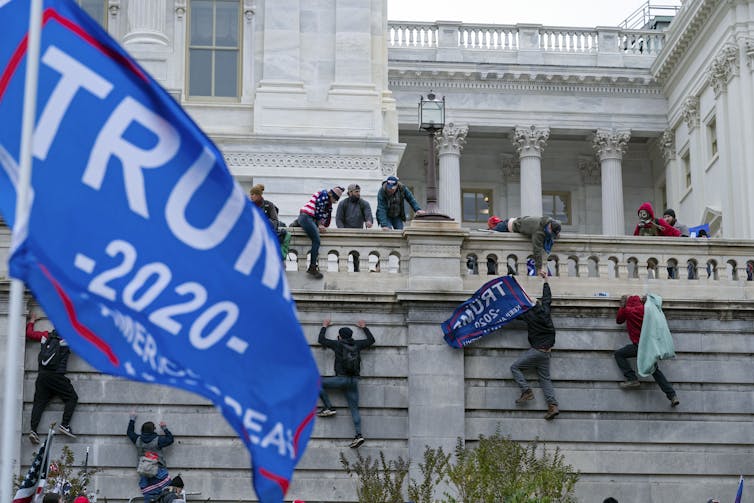 Rioters climb a wall into the U.S. Capitol with a Trump flag in the foreground.