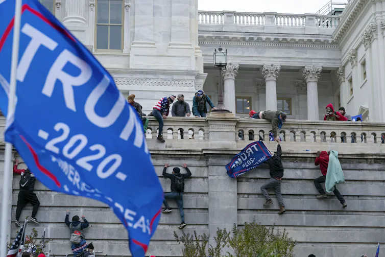Rioters climb a wall into the U.S. Capitol with a Trump flag in the foreground.