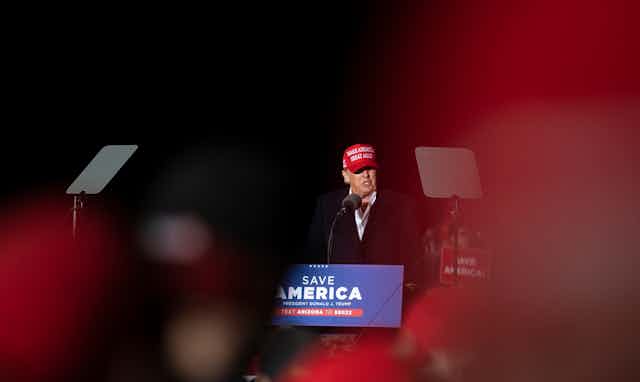 A man in a red Make America Great Again speaks into a microphone in front of a Save America banner