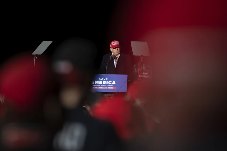 A man in a red Make America Great Again speaks into a microphone in front of a Save America banner