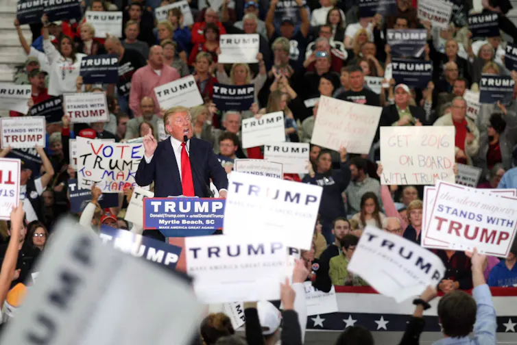 A man in a dark suit and red tie speaks at a rally of white people.