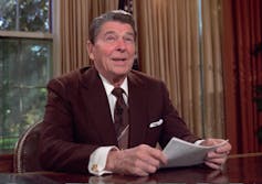 A man in a brown suit smiles at a desk with papers in his hands.