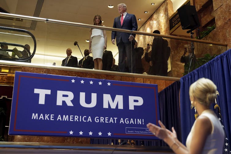 A man in a suit and a woman in a white suit stand at the the top of an escalator as another woman in a white dress looks up and applauds them.