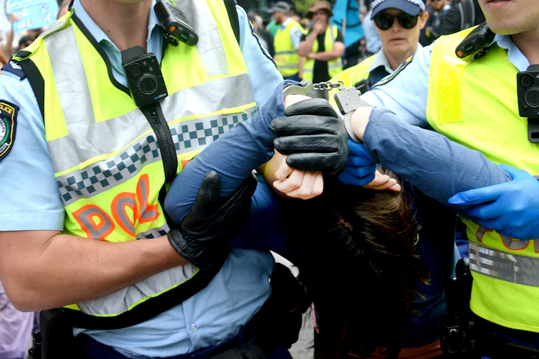 A person is being escorted away by police. Their hands are in handcuffs and their face is covered.