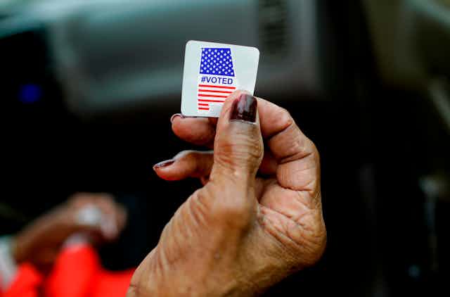 A Black voter's hand holds a #voted sticker.