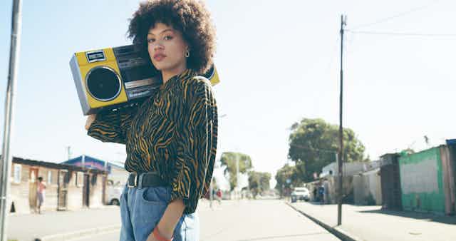 A young woman stands in the street of a working class suburb. She holds a radio to her shoulder, wears an Afro hairstyle and blue jeans with an animal print blouse.