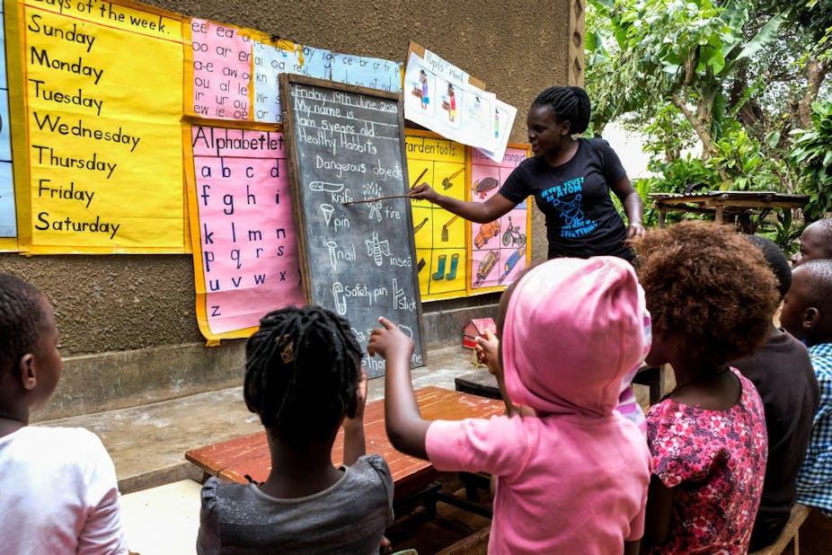 A female kindergarten teacher teaching children in an outdoor area. She's pointing at a blackboard propped against the wall with educational posters behind it.