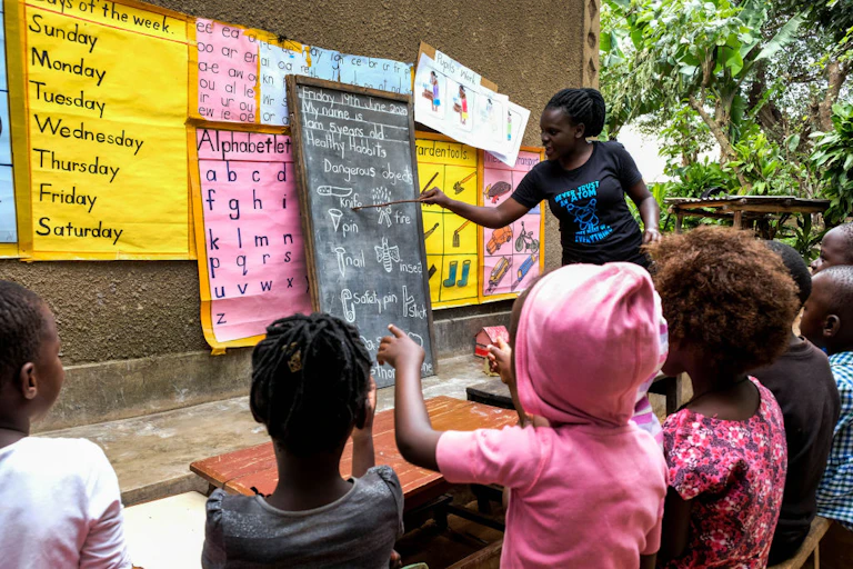 A female kindergarten teacher teaching children in an outdoor area. She's pointing at a blackboard propped against the wall with educational posters behind it.
