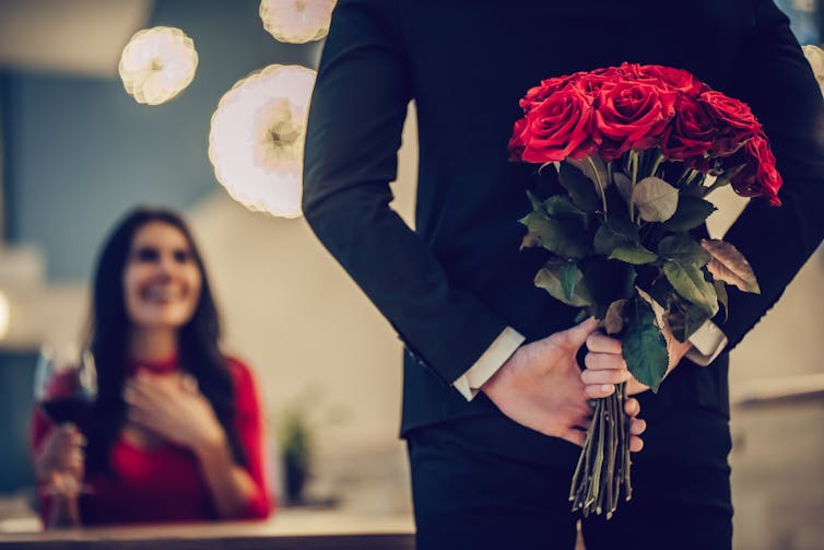 A man with roses hidden behind his back approaches a smiling woman .