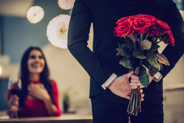 A man with roses hidden behind his back approaches a smiling woman .