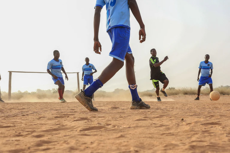 A dusty football field with a group of young footballers kicking the ball, among them an older player in a black football outfit.