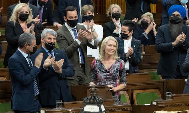 A blonde woman in a flowered dress stands in the House of Commons surrounded by people in masks applauding her.