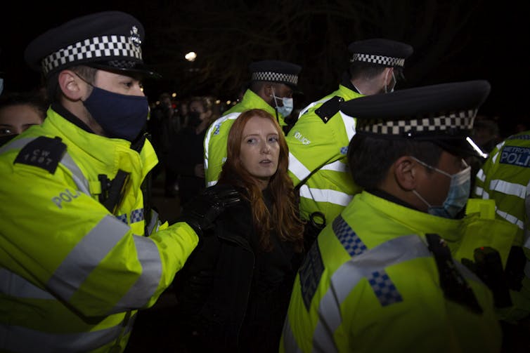 A woman is flanked by five police officers as she is arrested.