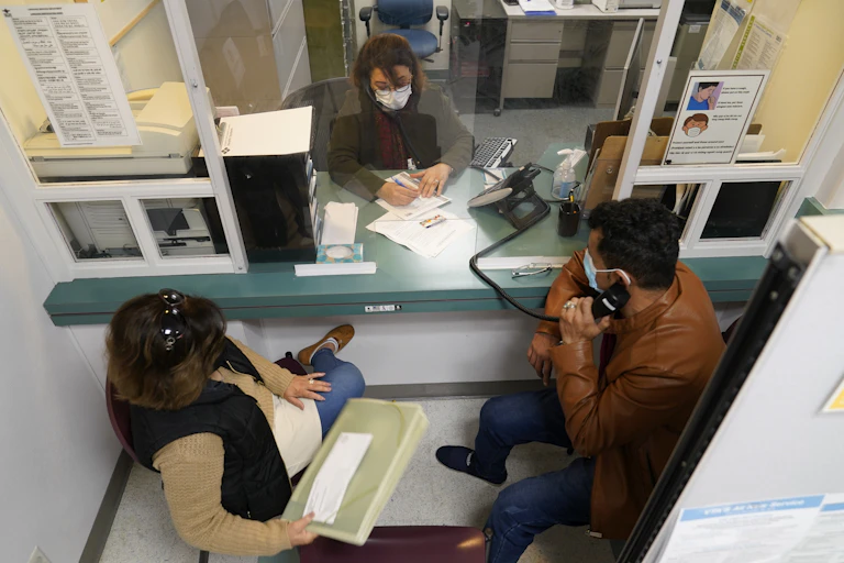 A man and woman sit at a reception desk opposite a woman with paperwork as the man holds a telephone to his ear.