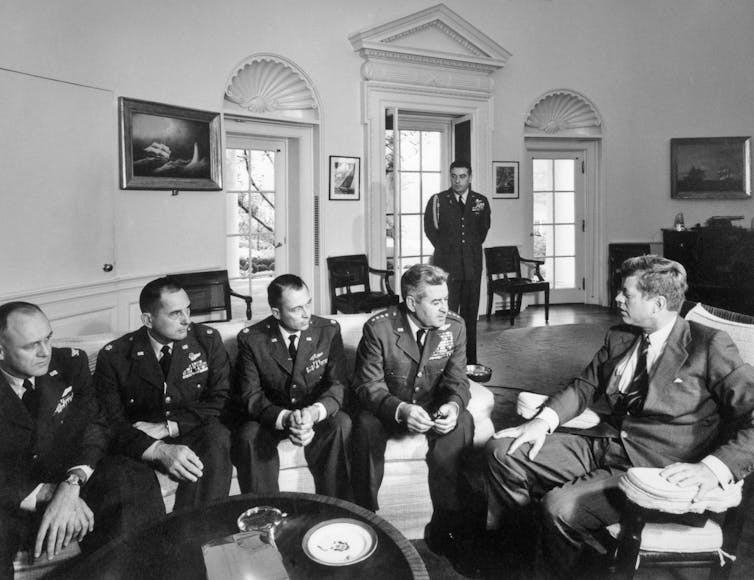 A group of men in military uniforms meets in an office with another man.