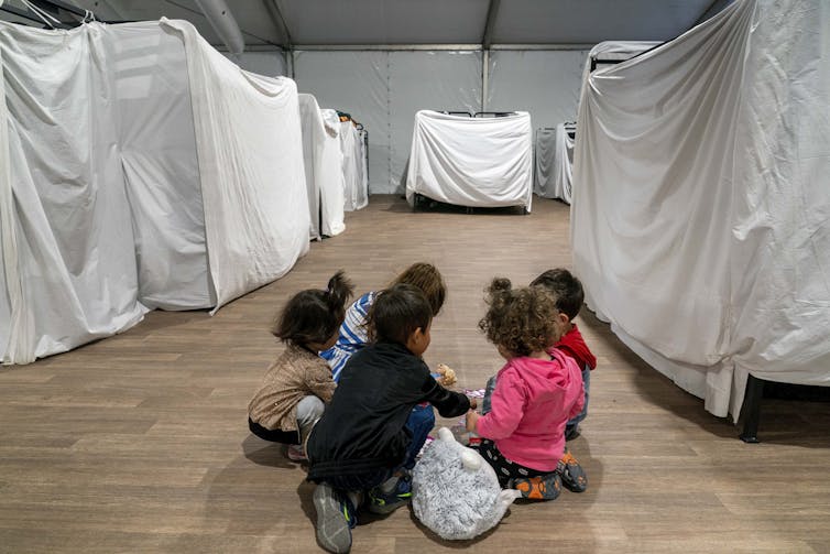 Several seated young children play on a bare floor surrounded by metal frames draped in white tarps.
