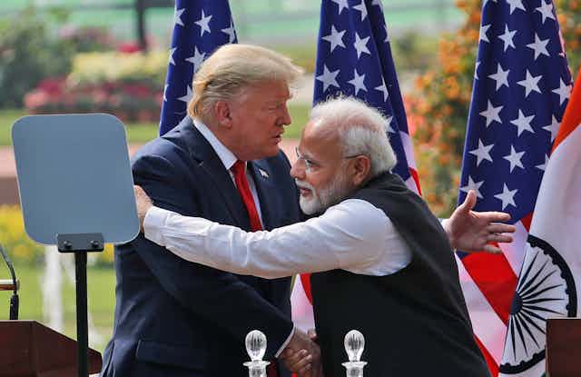 Modi embraces Trump. Behind them, stand three American flags. 