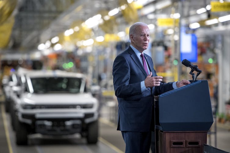 Biden at a lectern with Hummer EVs in the assembly line behind him.