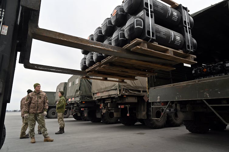 Soldiers watch a forklift load a crate of weapons onto the back of a truck.