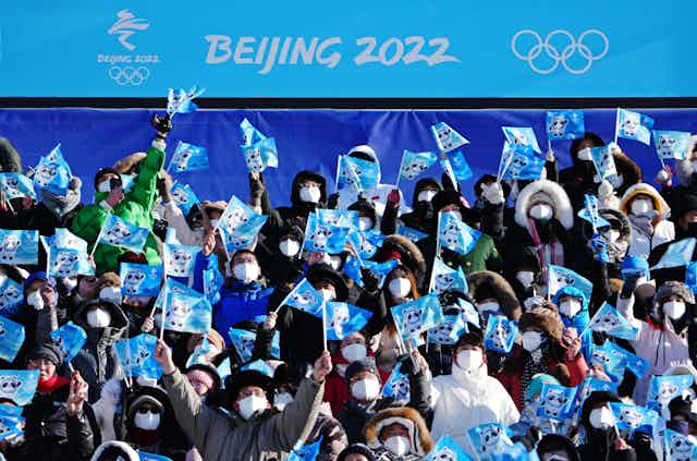 People in the stands wearing masks are waving small flags as they watch the snowboarding competition.