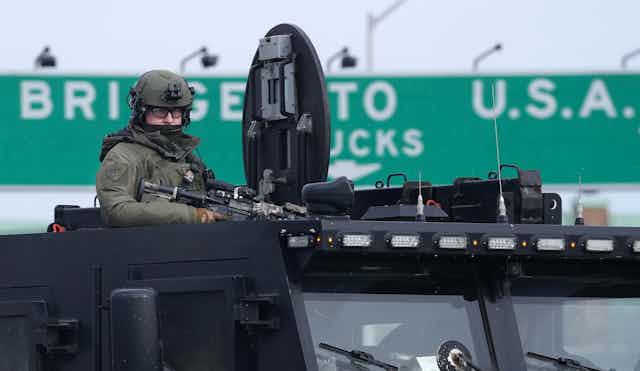 A police officer in tactical gear and holding a weapon at the top hatch of an armoured vehicle.