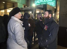 A bearded man in a dark jackets talks to a woman in a grey coat outside a venue.