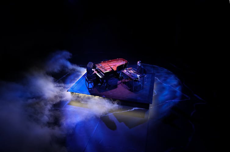 A woman plays piano, a man sits at a desk.