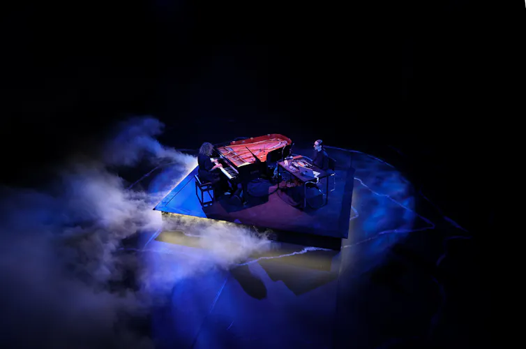 A woman plays piano, a man sits at a desk.