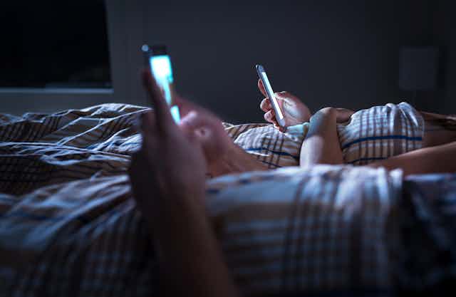 Two people in bed lazily scroll through their phones at night