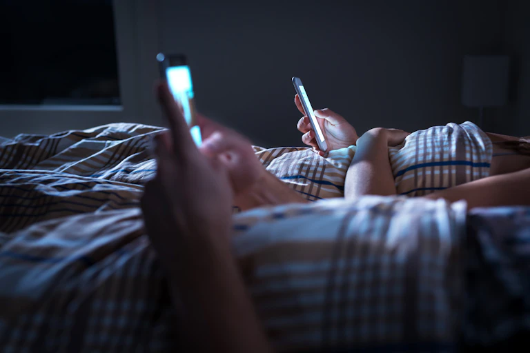 Two people in bed lazily scroll through their phones at night