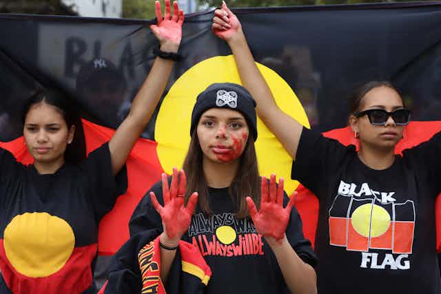 three women with red paint on hands and Aboriginal flag